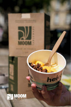 Charger l'image dans la galerie, Person holding a cup of dessert with a wooden spoon, featuring MOOM packaging in the background, emphasizing sustainable tableware.