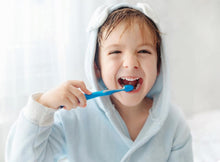 Charger l'image dans la galerie, Child brushing teeth with blue toothbrush, smiling in cozy bathrobe, promoting healthy dental hygiene.