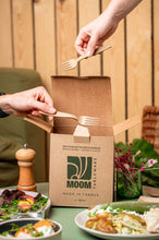 Charger l'image dans la galerie, Two hands placing reusable 16cm forks into a kraft box labeled "MOOM Made in France" with food items on the table.