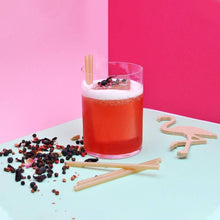 Charger l'image dans la galerie, Cocktail served with bagasse straws on a colorful background with scattered herbs and a wooden flamingo decoration.